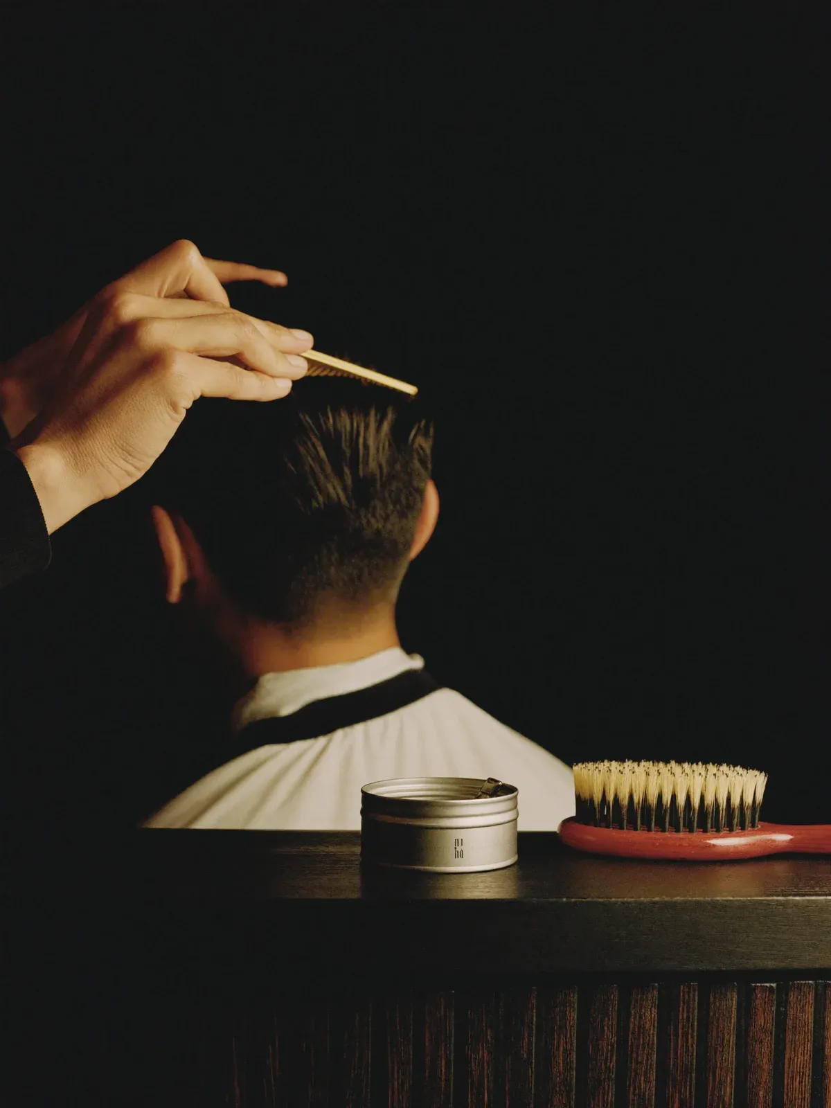 Barbier kamt het haar van een klant in model; een geopende pommadedoos en een borstel staan op het donkere blad